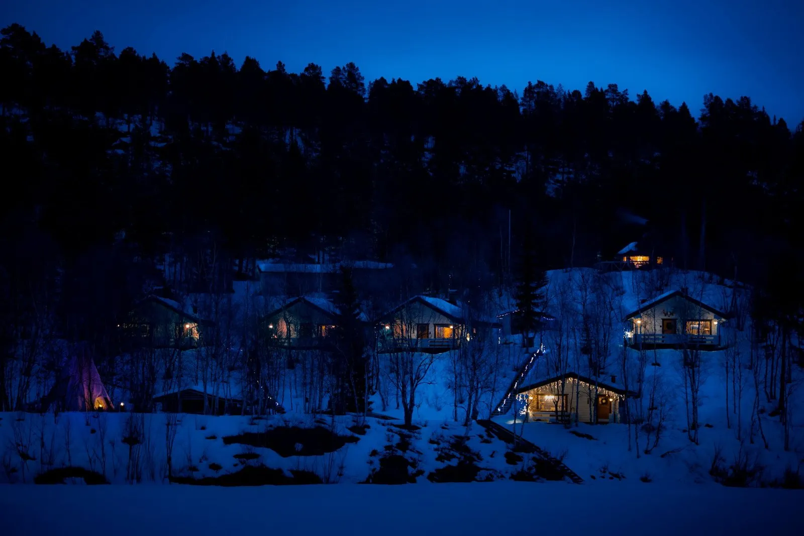 Napakettu Holiday Villagen mökit valaistuina lumisella rinteellä Inarijoen rannalla